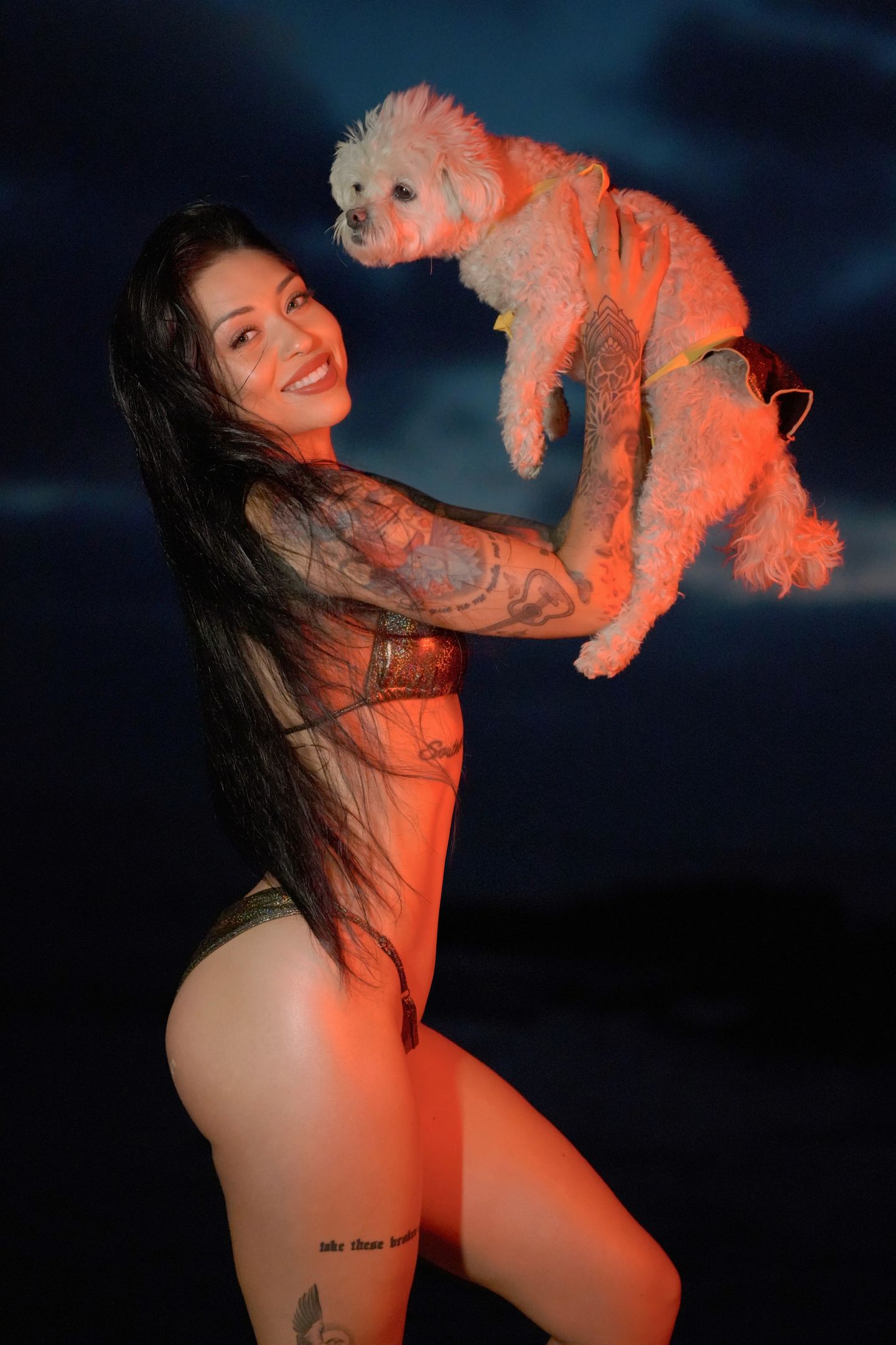 A smiling woman with long dark hair and tattoos wearing a bikini holds a small white dog up against a dark night sky.