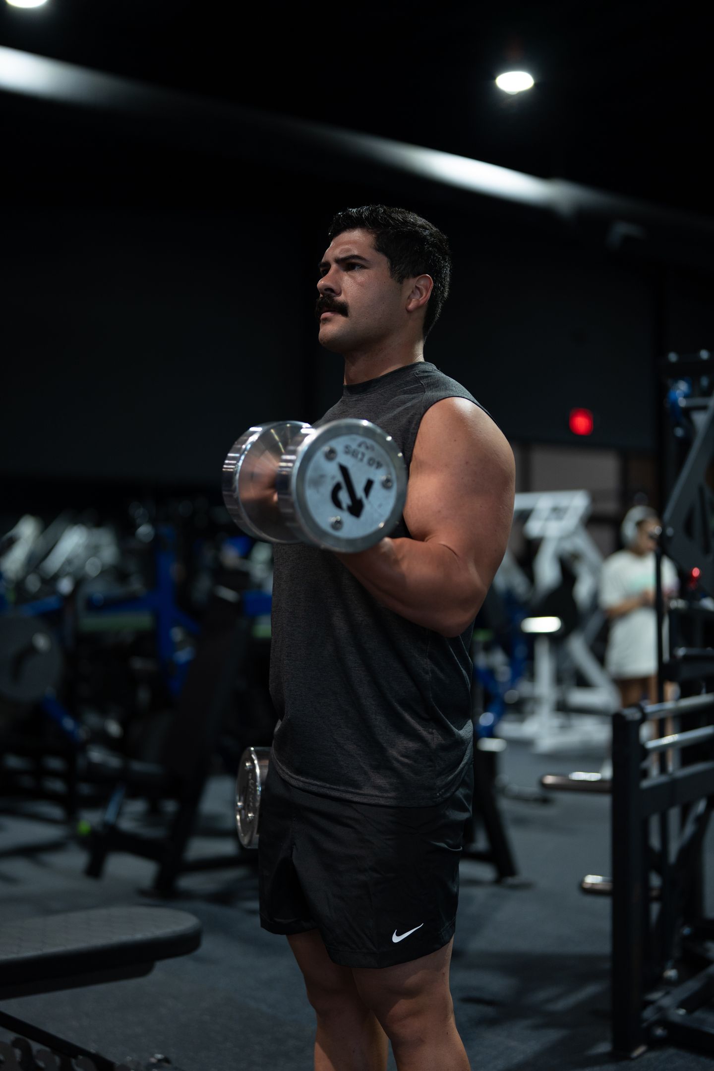 A man with a mustache performs a bicep curl with a dumbbell in a dimly lit gym.