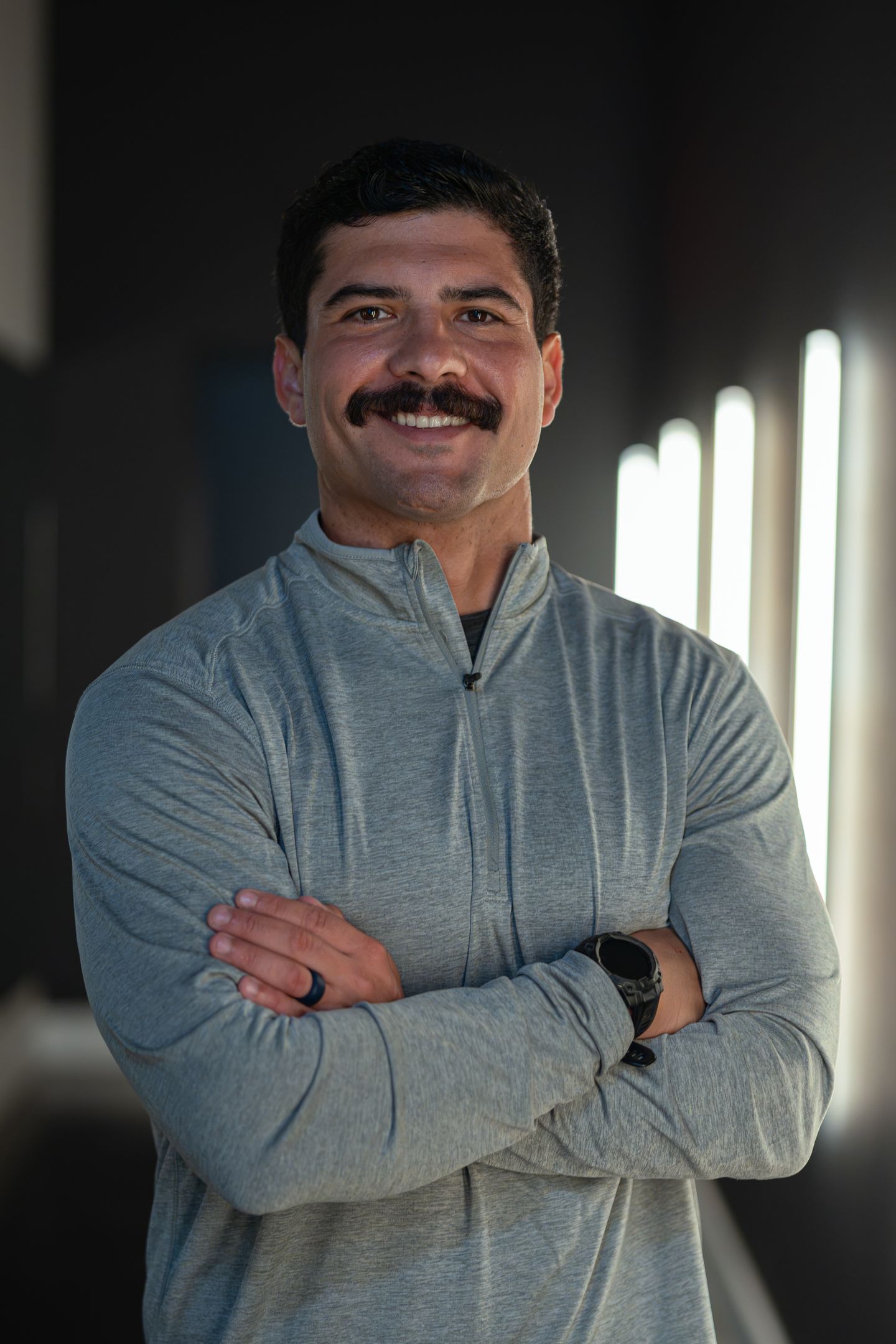 A smiling man with a mustache stands with his arms crossed in a gym setting with vertical lights in the background.