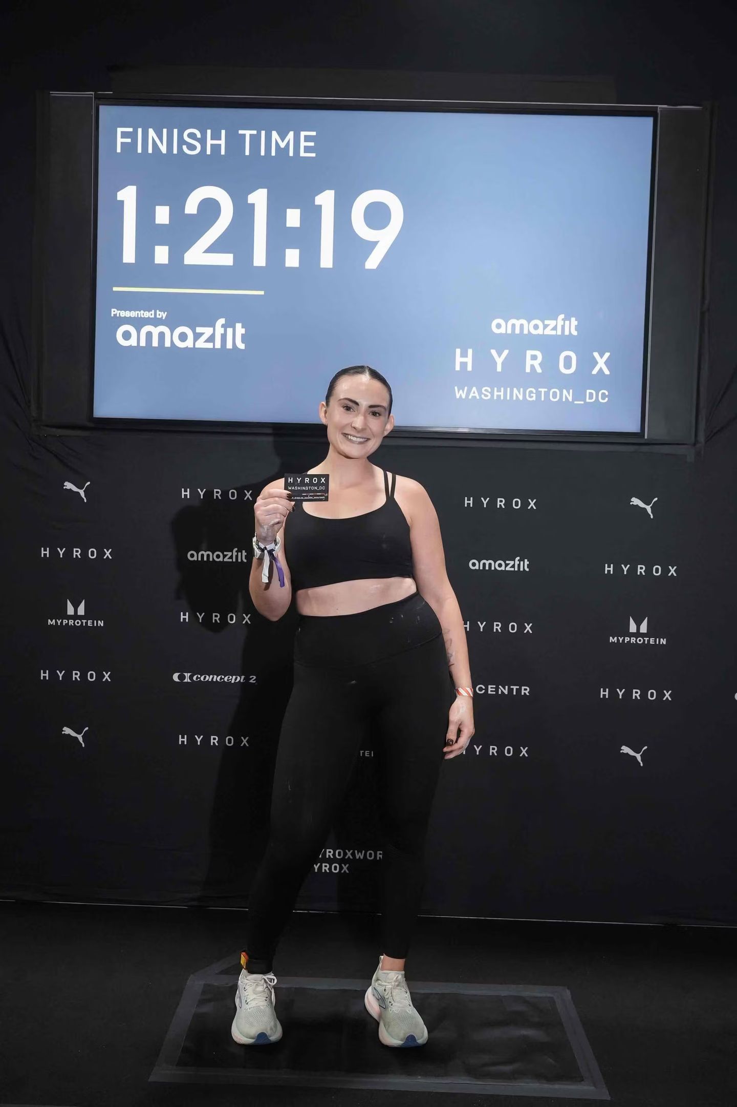 A smiling woman in athletic wear holds a race medal in front of a screen displaying her finish time of 1:21:19.
