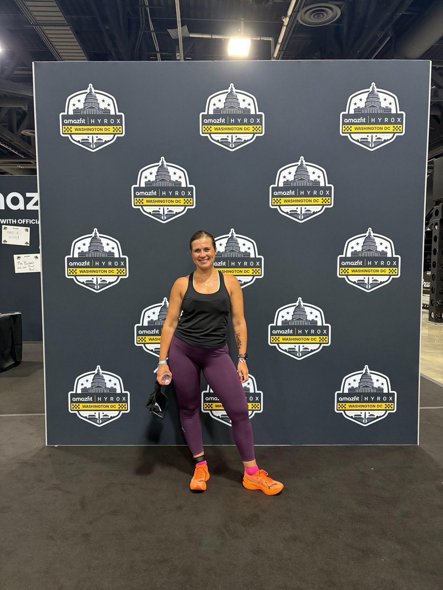 A woman in athletic wear stands smiling in front of a branded event backdrop.