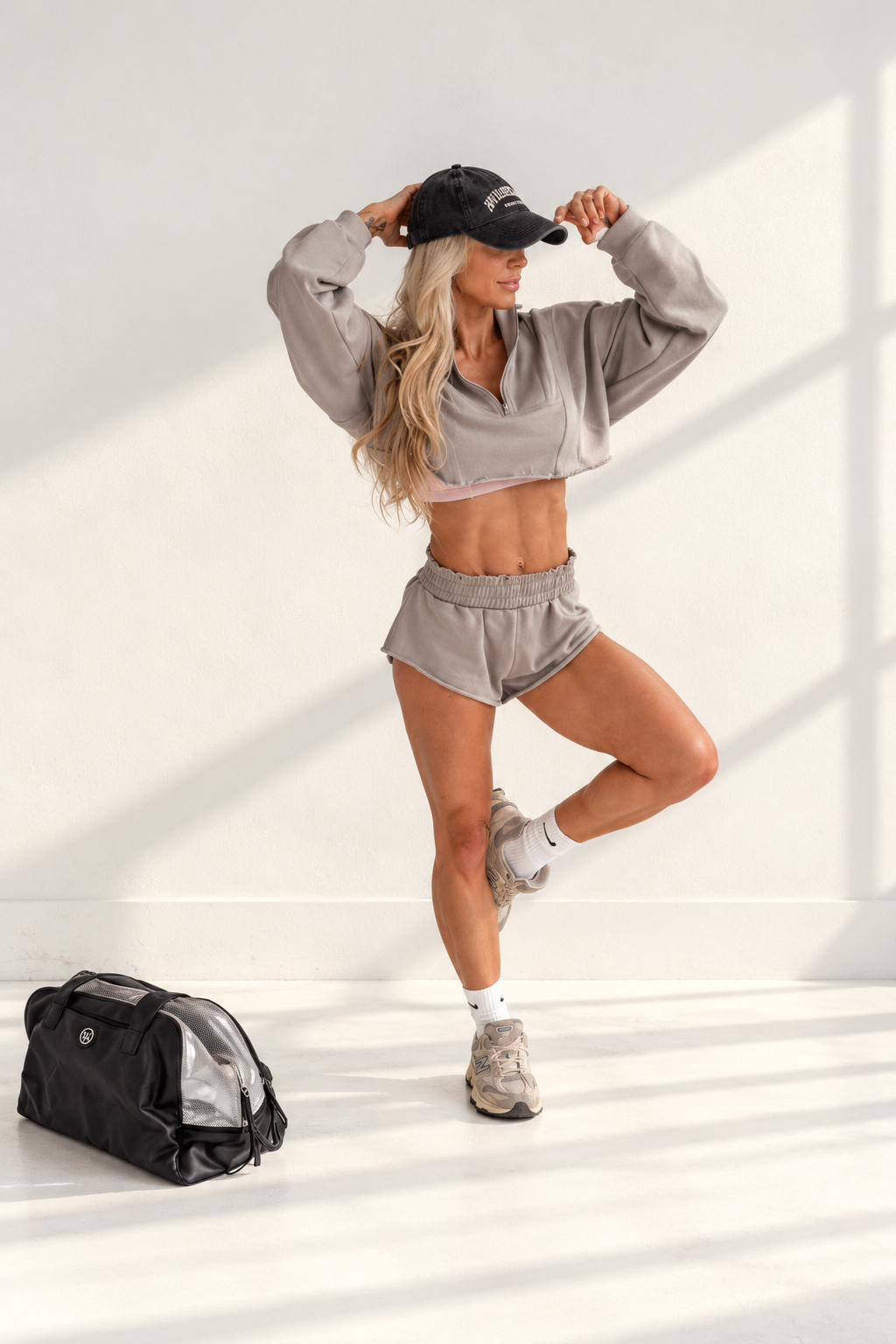 A fit woman in a cropped sweatshirt and shorts poses with her hands on her cap next to a gym bag in a bright studio.