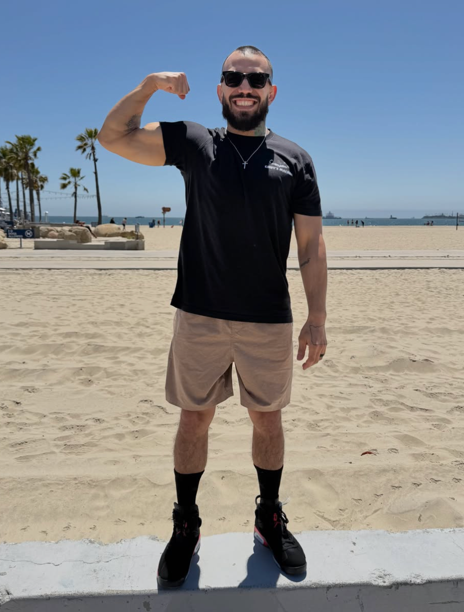 A smiling man with a beard and sunglasses flexes his bicep while standing on a sunny beach.