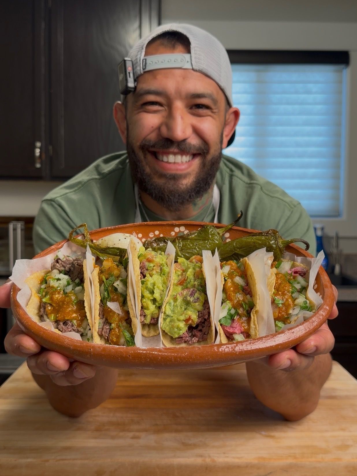A smiling man holds a large clay platter filled with several tacos topped with guacamole and salsa in a kitchen.
