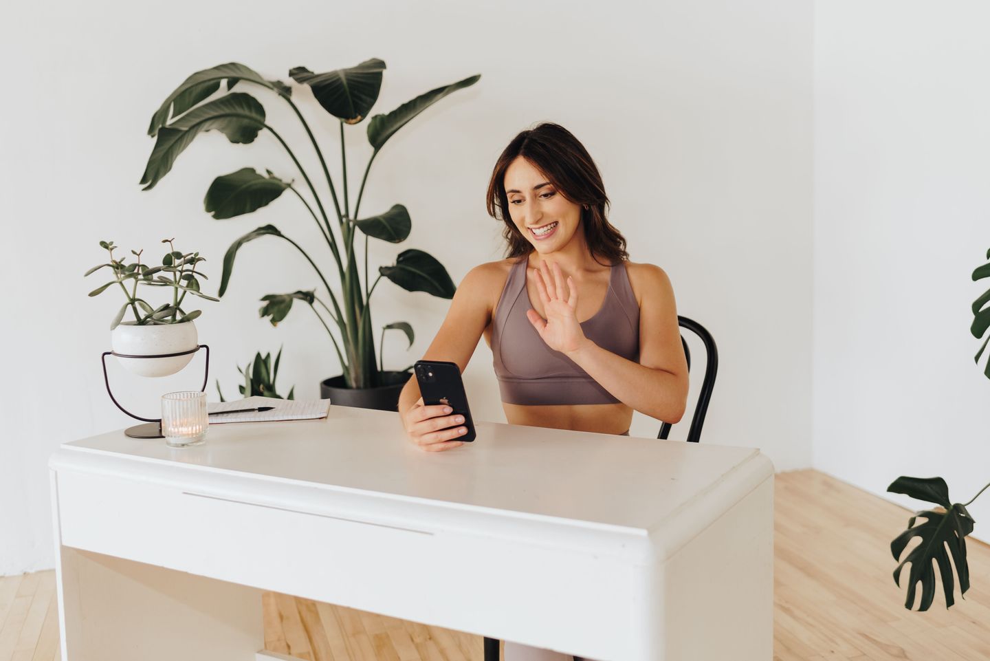 A woman in athletic wear sits at a desk and waves while on a video call on her smartphone.