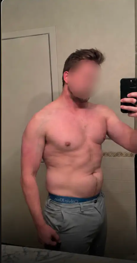 A shirtless man takes a mirror selfie in a bathroom to show his physique.