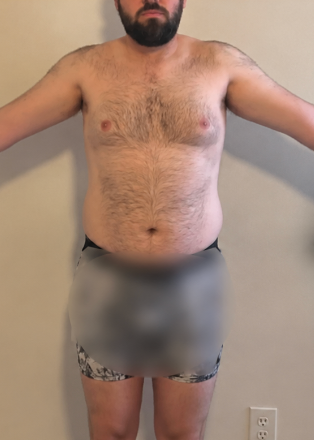 A shirtless man with a beard stands facing forward against a plain wall for a fitness progress check.