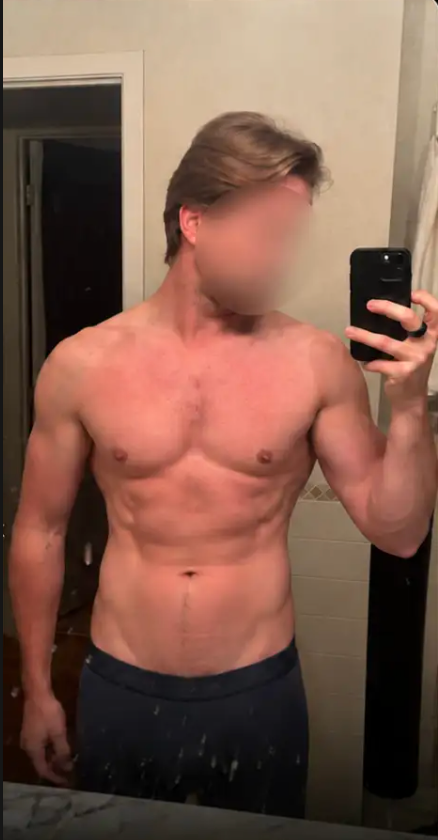 A shirtless man with a muscular build takes a mirror selfie in a bathroom.