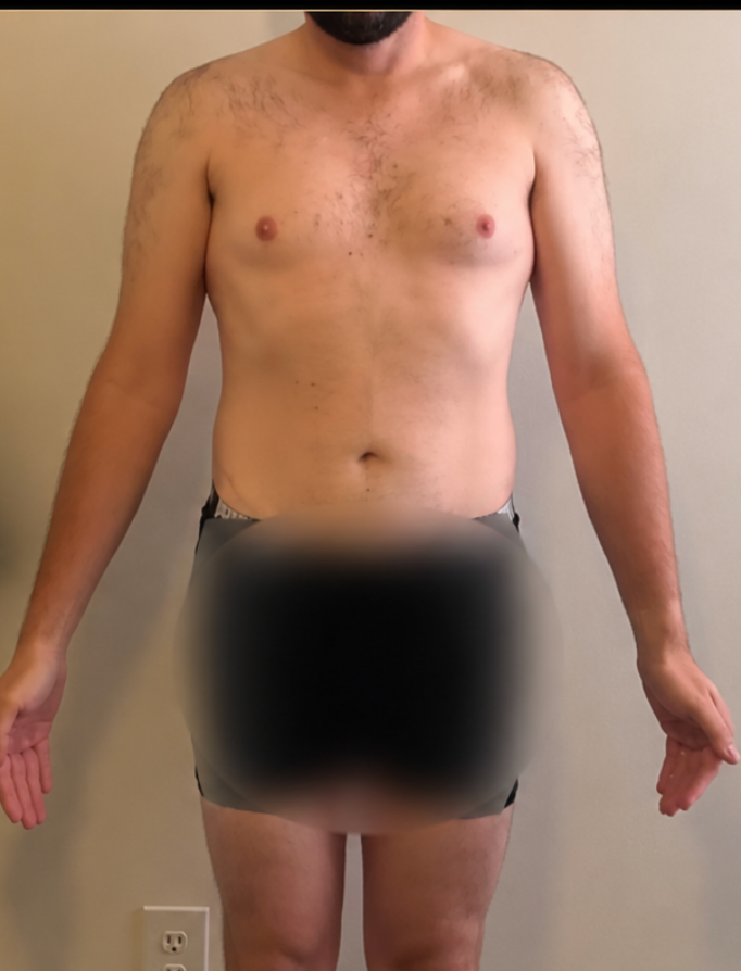 A man stands shirtless against a plain wall for a fitness progress check.