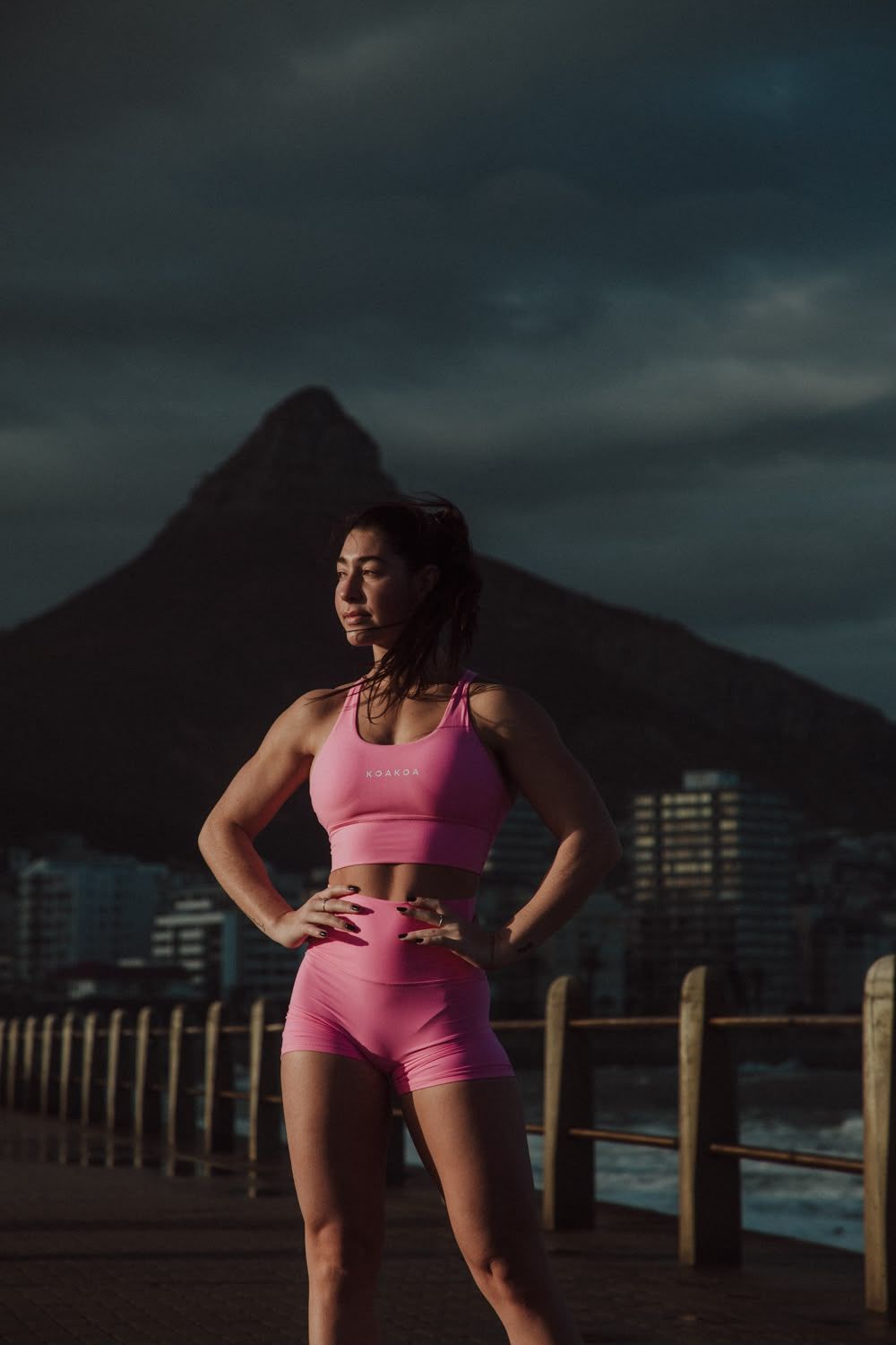 A woman in pink athletic wear stands with hands on her hips on a boardwalk with a mountain in the background.