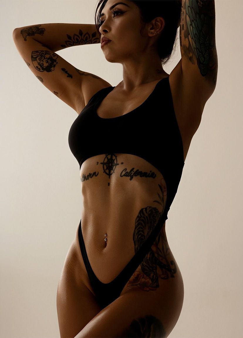 A fit woman with tattoos poses with her arms raised, showcasing her toned physique in a black athletic outfit.