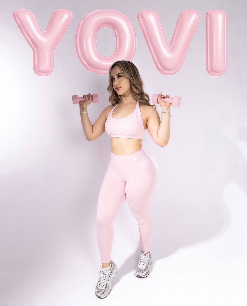A woman in pink activewear holds small dumbbells in front of large pink balloons spelling out Yovi.