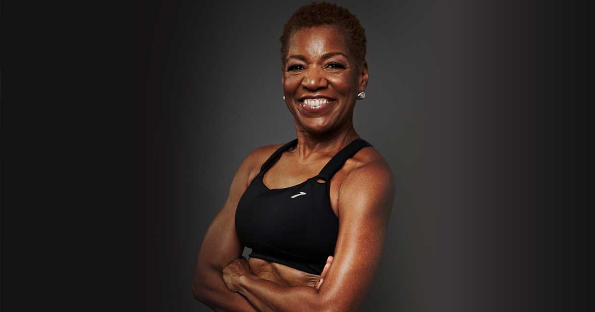 A smiling woman with short hair wearing a black sports bra stands with her arms crossed against a dark background.