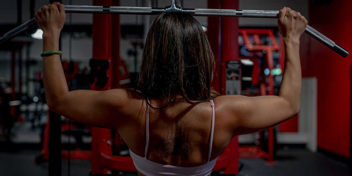 A woman seen from behind performs a lat pulldown exercise on a machine in a gym.