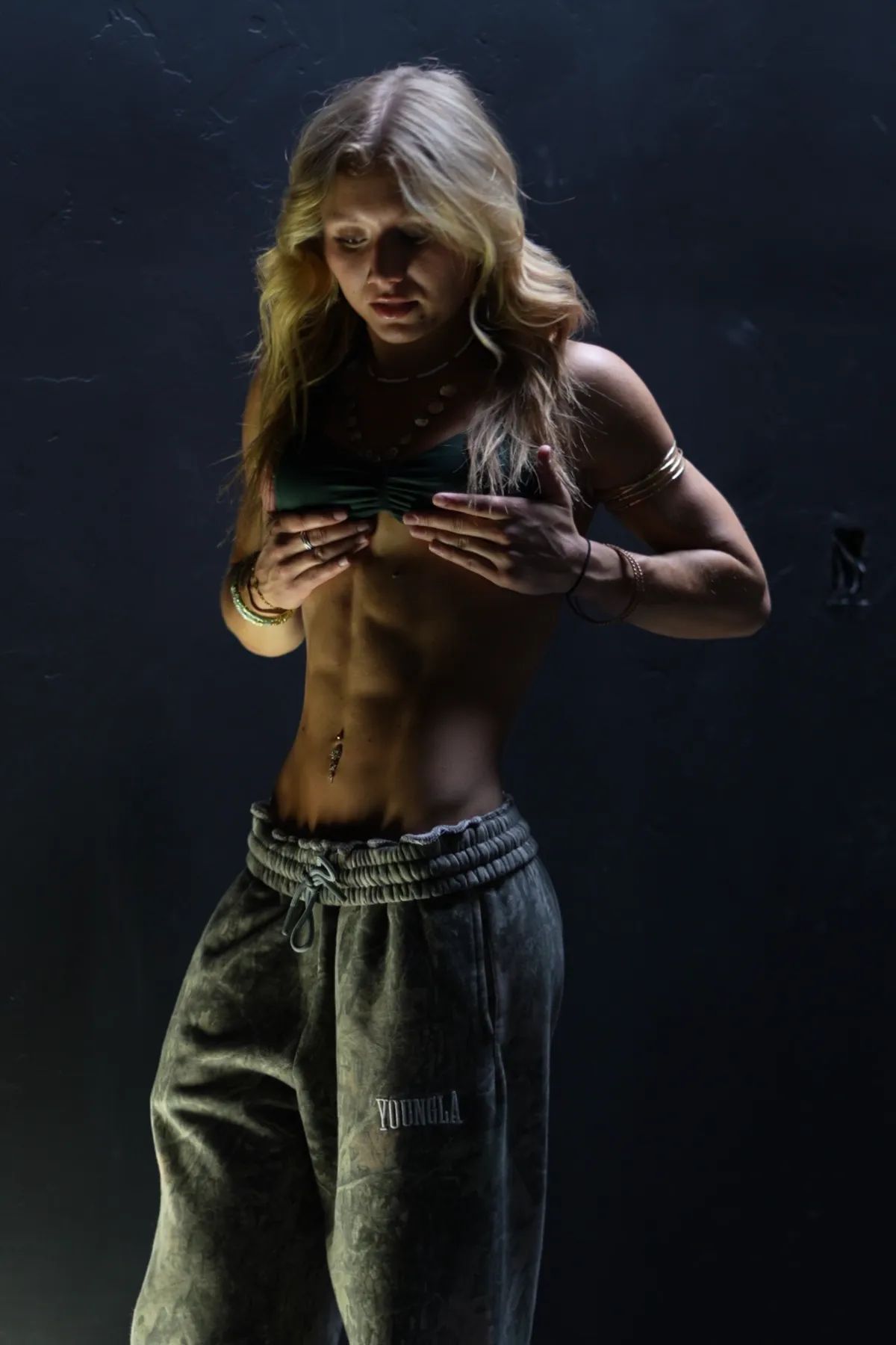 The coach showing her defined abs in a dark room, wearing a top and baggy sweatpants.